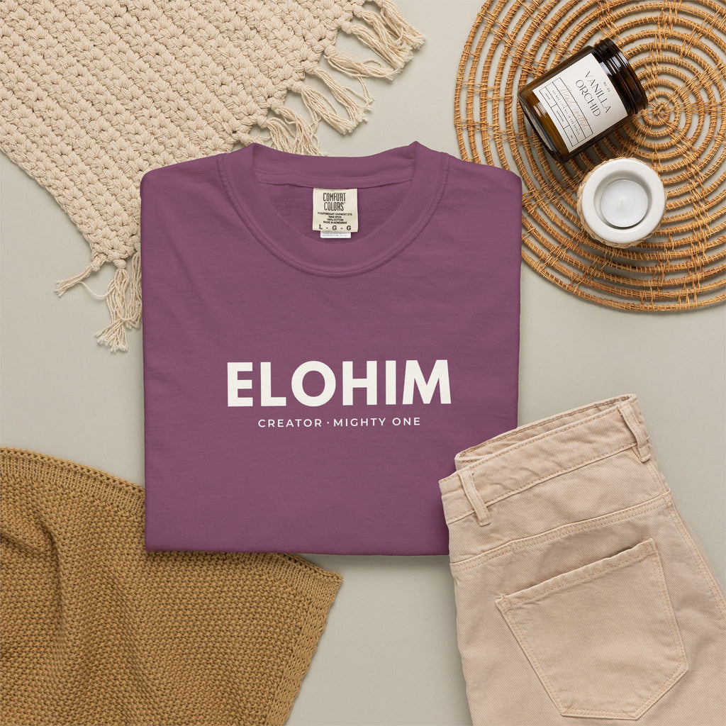 Berry purple Elohim Christian T-shirt with “Creator · Mighty One” typography, styled in a neutral flat lay.