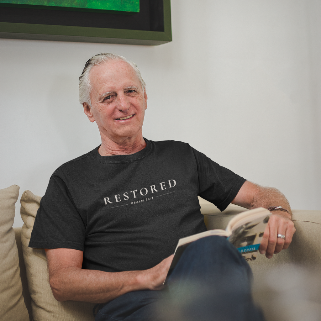 Man wearing black Restored Christian T-shirt with Psalm 23:3 typography while seated indoors