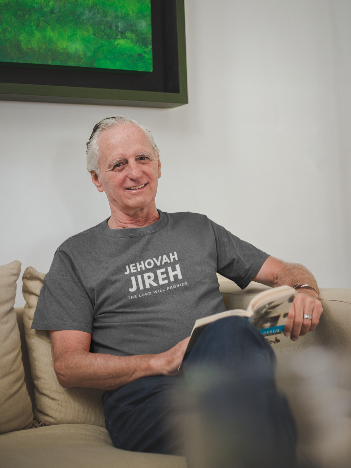 Older man reading on a sofa while wearing a gray Jehovah Jireh Christian T-shirt with “The Lord Will Provide” printed in white.